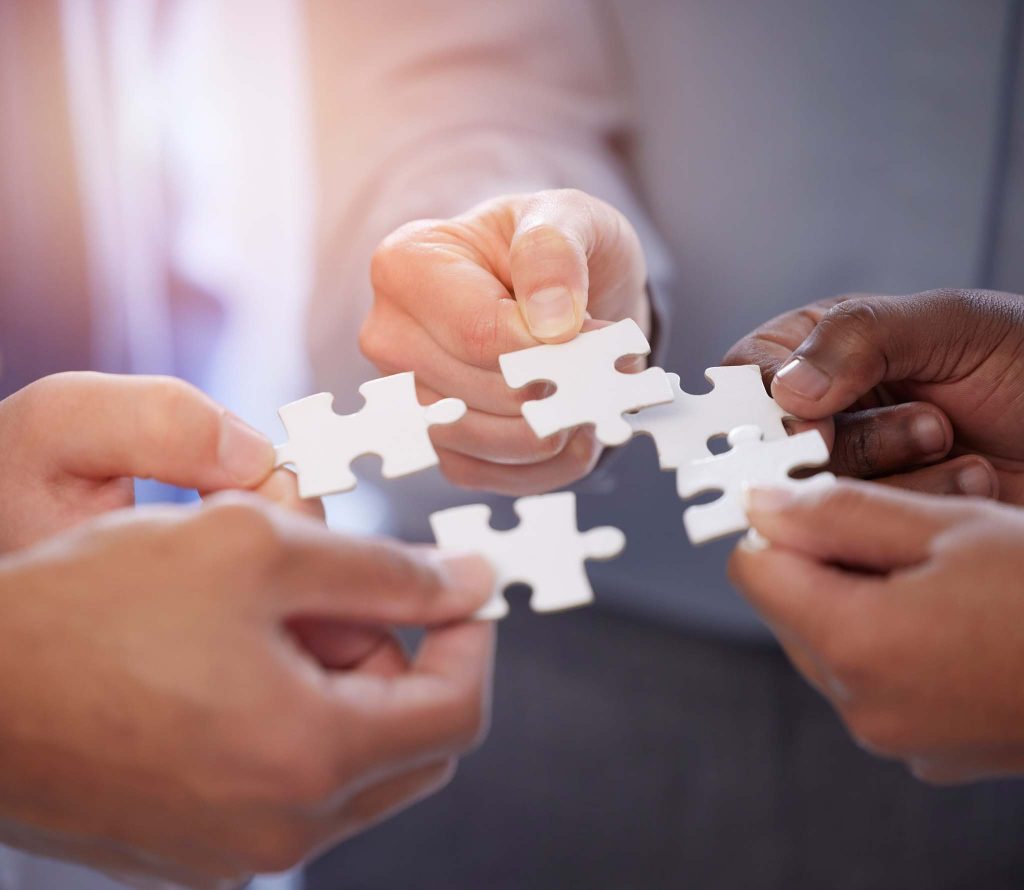 Een close-up van verschillende handen die samen puzzelstukjes in elkaar leggen op een tafel. De afbeelding weerspiegelt het idee van het bundelen van keuringen door VvE’s, pandeigenaren en huurders om tijd, kosten en inspanningen te delen.